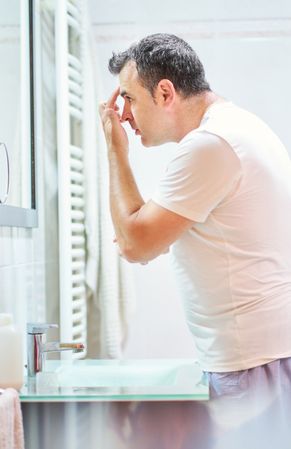 Man touching face in bathroom while looking in mirror