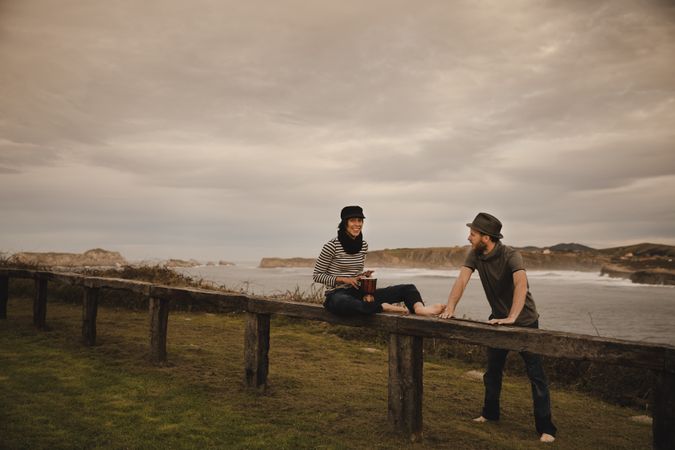 Young guy in hat near elegant woman playing on hand drum in cap on seat near coast of sea and cloudy sky