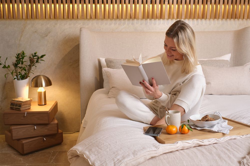 Young female having breakfast and reading book in bedroom