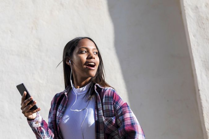 Happy female standing in the sun in front of wall singing along with music on phone