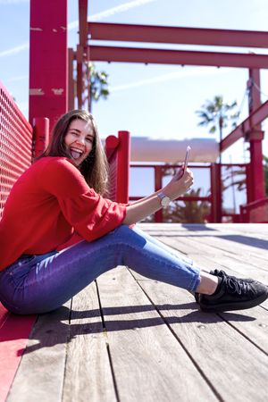 Side view of happy beautiful young woman sitting on ground and looking at camera while using a mobile phone