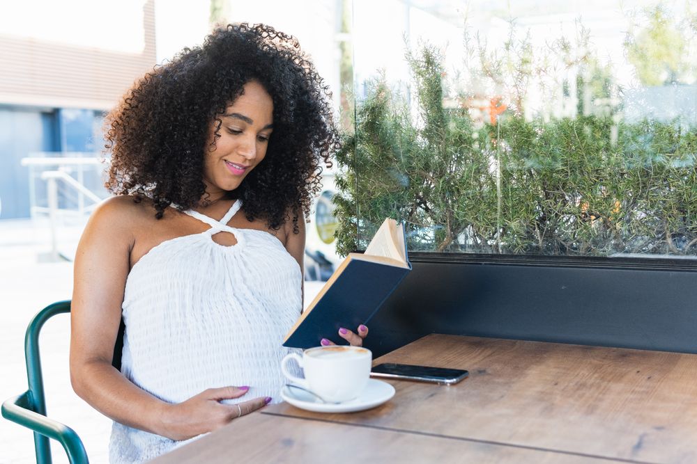 Pregnant Black woman reading book and drinking coffee