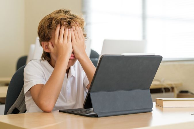 Frustrated schoolboy covering face at table in classroom