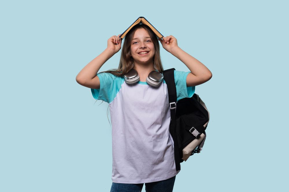 Playful girl with book on head, wearing backpack and headphones