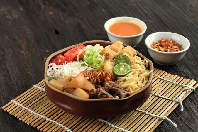 Indonesian beef noodle soup with noodles, beef, spring roll, cabbage and tomato served on mat
