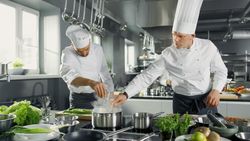 Two Famous Chefs Work as a Team in a Big Restaurant Kitchen. Vegetables and Ingredients are Everywhere, Kitchen Looks Modern with Lots of Stainless Steel.