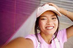 Happy Chinese female looking up at camera while taking a selfie of herself next to pink wall outside