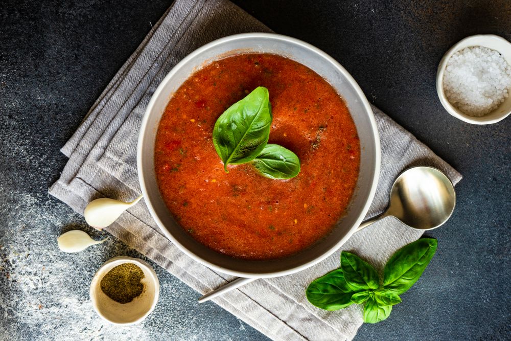 Top view of grey bowl of gazpacho soup with basil leaves on napkin served with seasoning