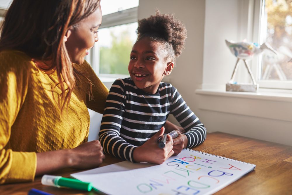 Black mother and daughter smile at each other as they write the alphabet at home