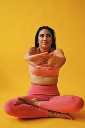 Hispanic female sitting in yoga pose stretching her arms in front of her in yellow studio, vertical