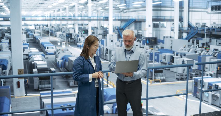 Factory Managers Working on Laptop at a Modern Manufacture with Autonomous Robot Hands. Female and Male Specialists Monitoring Technical Data on a Computer, Analyzing Reports and Planning Updates