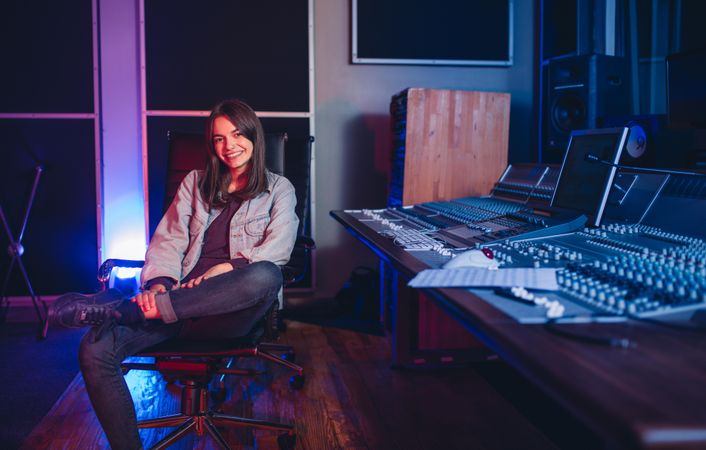 Happy young woman sitting in music studio next to sound board