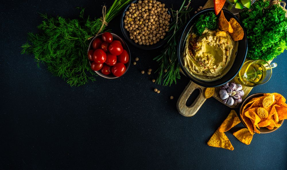 Top view of creamy hummus dip in bowl on board served with olive oil, veggies and chick peas