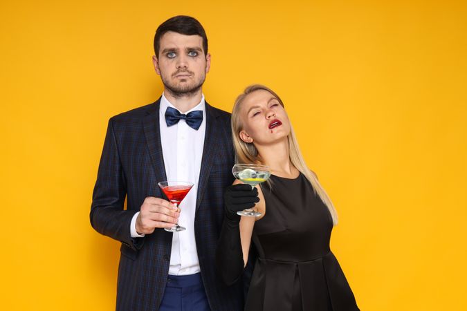 A couple in Halloween costumes on a yellow background with cocktails in their hands