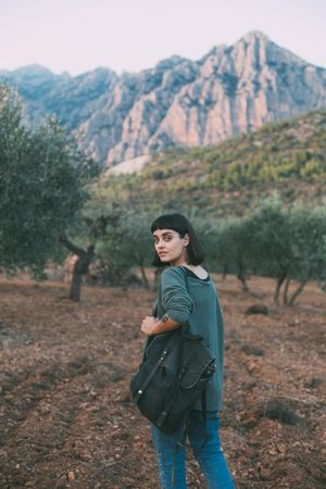 Woman with large book bag on shoulder looking around park with olive trees