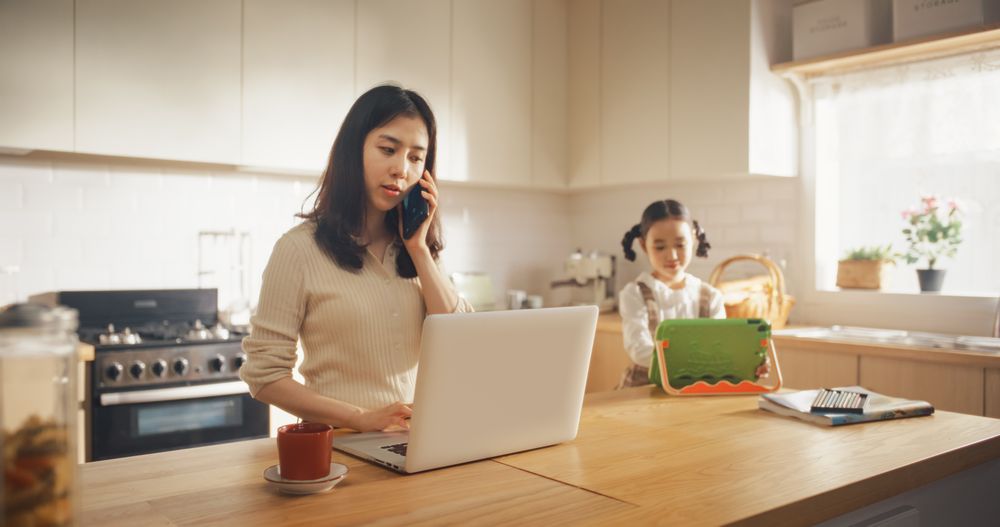 Portrait of Korean Mother Using Laptop and Smartphone To Work from Home While her Little Daughter is Using Digital Tablet.