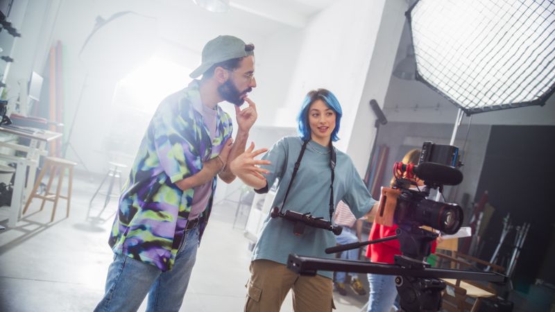 Young Female And Male Filmmakers Discuss Over A Camera Setup In A Bright Studio, Reflecting Teamwork And Creativity In Modern Film Production And Advertising.