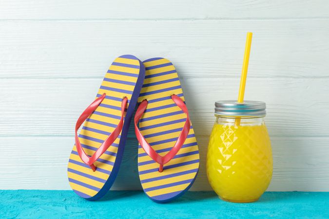 Flip flops and glass jar with fresh juice on color table against wooden background, space for text. Summer vacation backdrop