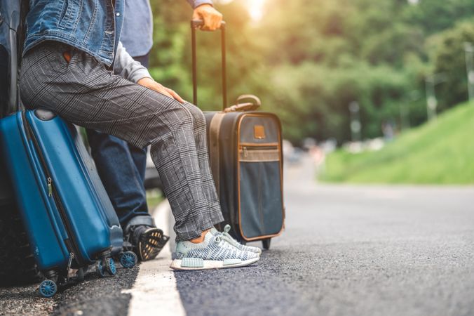 Two people in trainers with luggage sitting on side of road way, anonymous