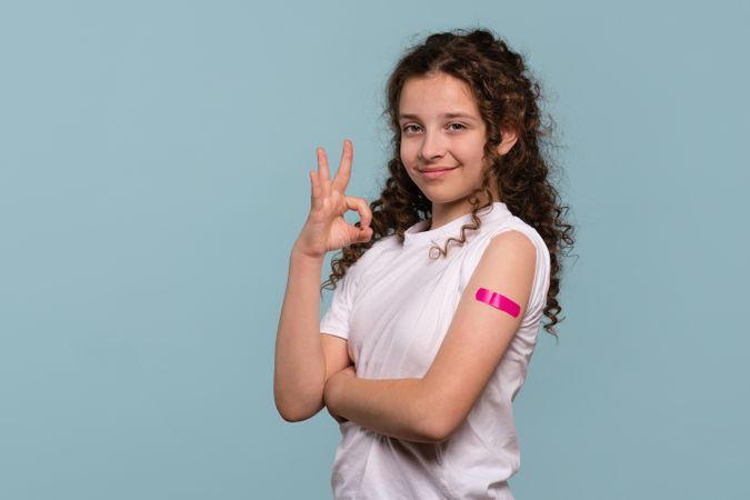 Girl making "OK" gesture with pink bandage on vaccinated arm.