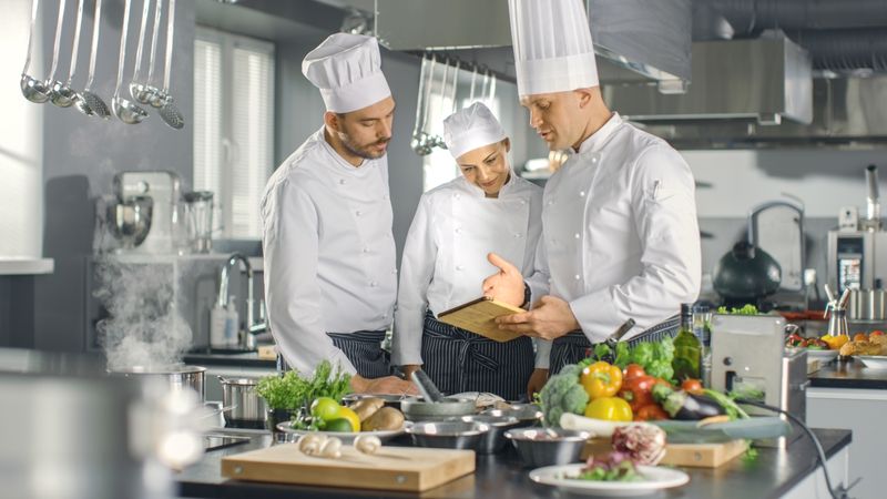 In the Modern Kitchen Team of Cooks Use Tablet Computer For Recipes, They Smile and Have Discussion. Kitchen is Full of Food Ingredients, Vegetables, Meat, Boiling Soup.