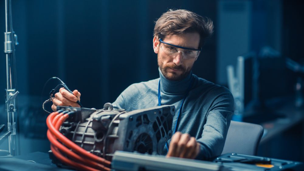 Professional Automotive Engineer in Glasses with a Computer and Inspection Tools is Testing an Used Electric Engine in a High Tech Laboratory with a Concept Car Chassis.
