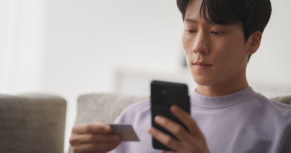 Portrait of a Stylish South Korean Man Online Shopping with His Smartphone While Sitting on a Couch in a Comfortable Living Room