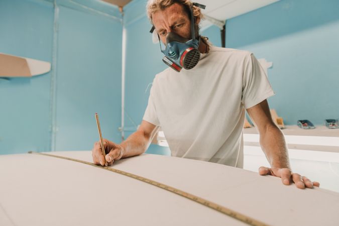 Man in respirator measuring surf board in workshop