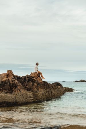 Lonely tourist sitting on rocky coast against tranquil sea water under gray sky