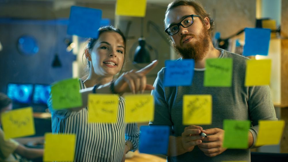 Male and Female Designers Discuss and Outline They're Project with Sticky Notes on a Glass Wall.