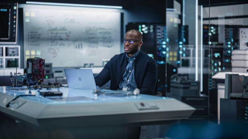 Portrait of Male Specialist Using Laptop Computer to Improve Electronic Circuit Board in Modern Office. Black Engineer Thinking and Typing Ideas for Artificial Intelligence Project