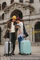 Two young women with suitcases smiling at tablet bxz3Z5