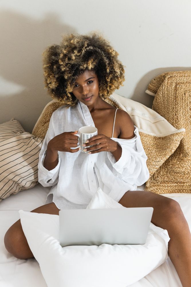 Black woman drinking coffee while sitting on bed with laptop
