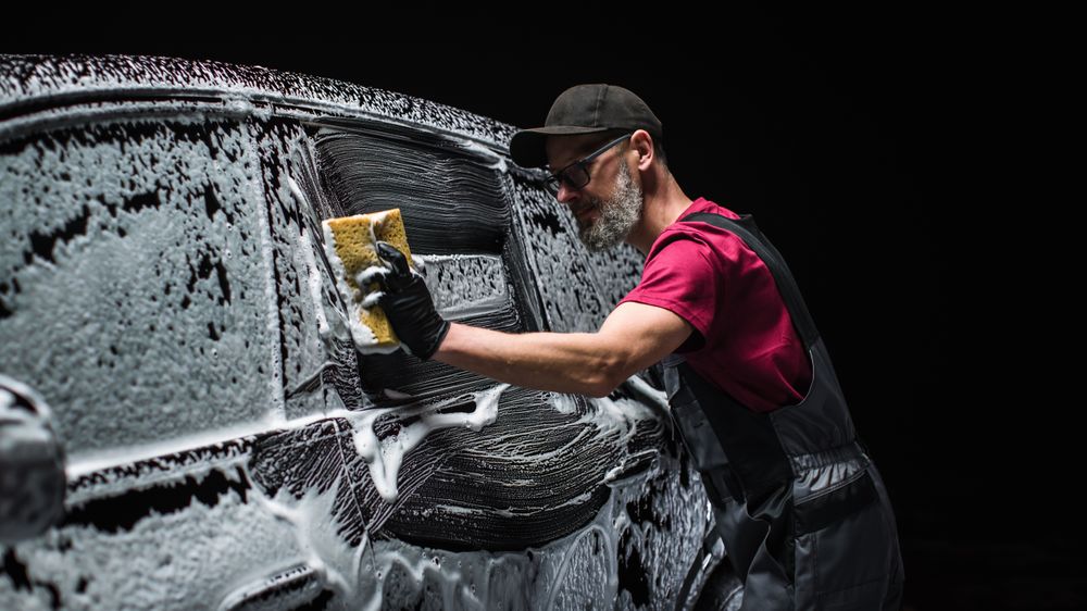 Portrait of a Professional Car Wash Specialist Using a Big Soft Sponge to Wash a High Tech Family Electric SUV with Foam Before Detailing,