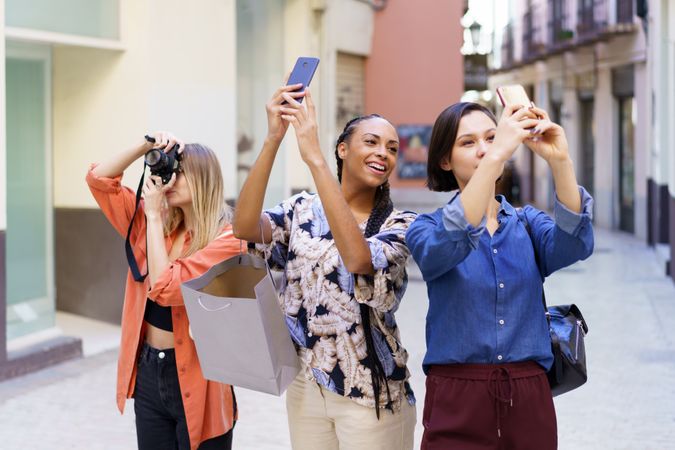 Three happy women looking up and taking pictures with their phone in narrow lane