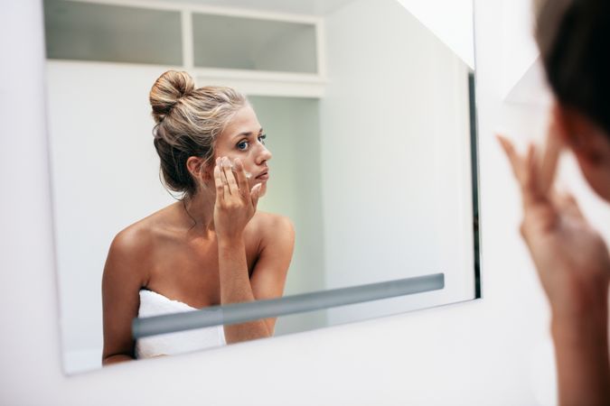Reflection of a woman in bathroom mirror applying cosmetic cream on her face