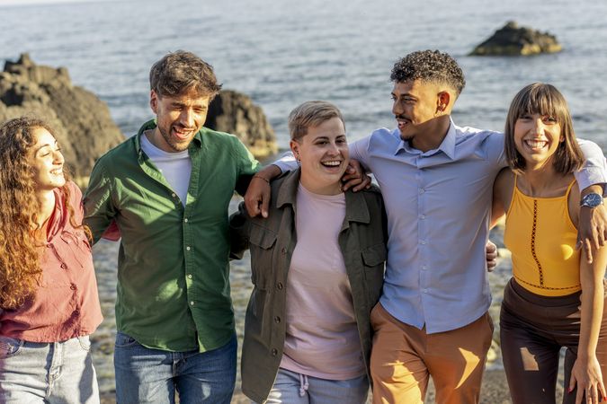Happy diverse group of friends hugging on rocky beach during golden hour