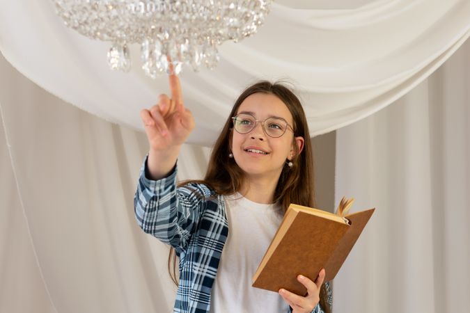 Teenage girl reading book touching crystal chandelier at home.