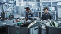 Indian And White Male Workers Taking Apart Laptops To Recycle For Production Of Digital Devices At Automated Electronics Factory With Robotic Arms.
