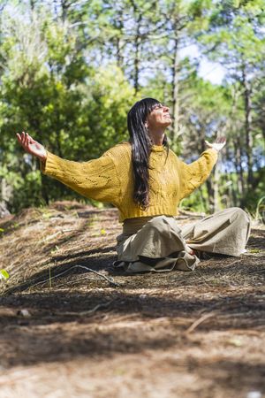 Woman sitting on the woods with arms opened while looking to the sky like thanking God. Vertical shot.