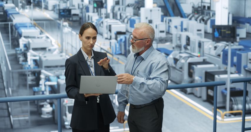 Experienced Male and Female Engineers Standing on a Platform at a Modern Industrial Factory, Using Laptop Computer and Discussing Production of Modern Electronics for Artificial Intelligence Machines