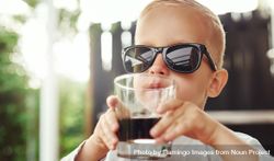 Blond boy in sunglasses sipping dark drink from glass outside bY9AYb