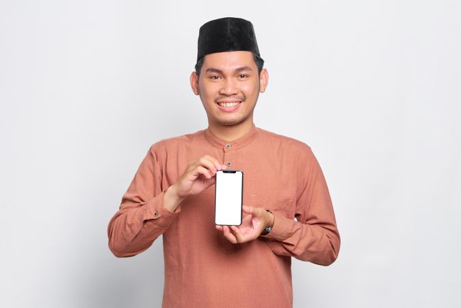 Happy Muslim man in kufi hat holding mobile phone with mock up screen with both hands