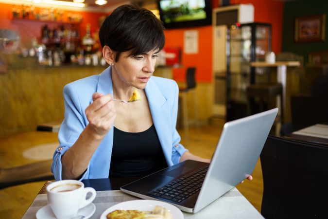Female in trendy blue jacket sitting at table using laptop while eating slice of cake in cafe