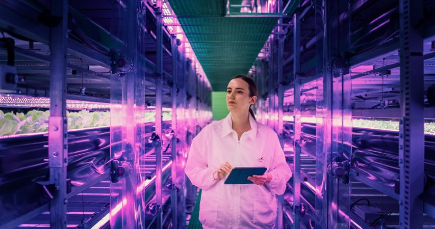 Female Hydroponics Specialist Walking in a Vertical Farm Corridor with LED Lights Illuminating the Plants.