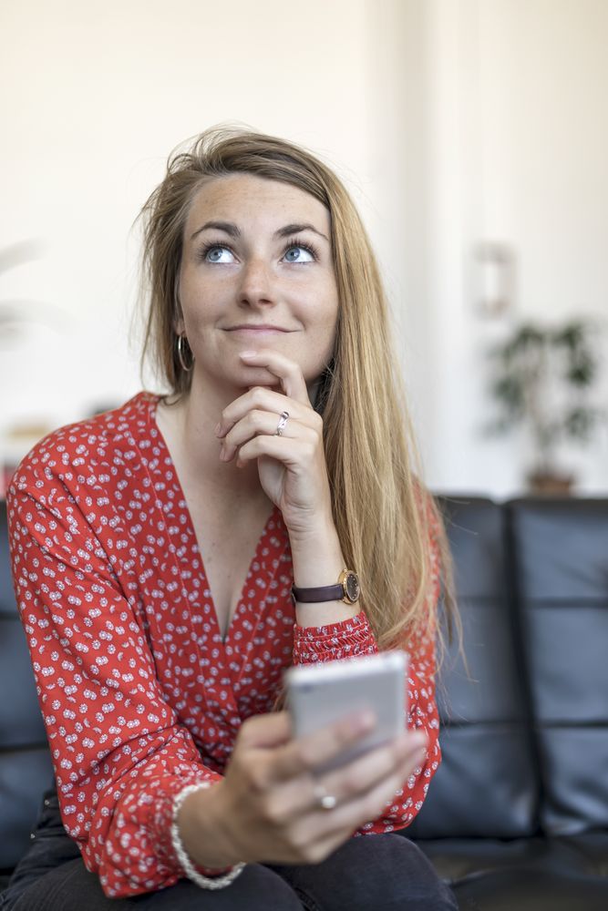 Portrait of white woman holding cellphone in hand and sitting on sofa while looking away thoughtfully