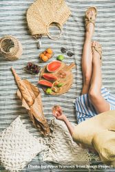 Woman on picnic blanket with  thatched bag with glass of rose, baguette, sliced watermelon and figs bDE1E0