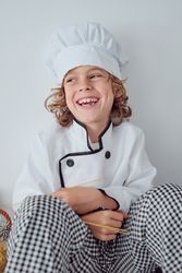 Cheerful boy in chef costume and cap near white wall