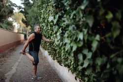 Mature man stretching leg holding botanical wall in street