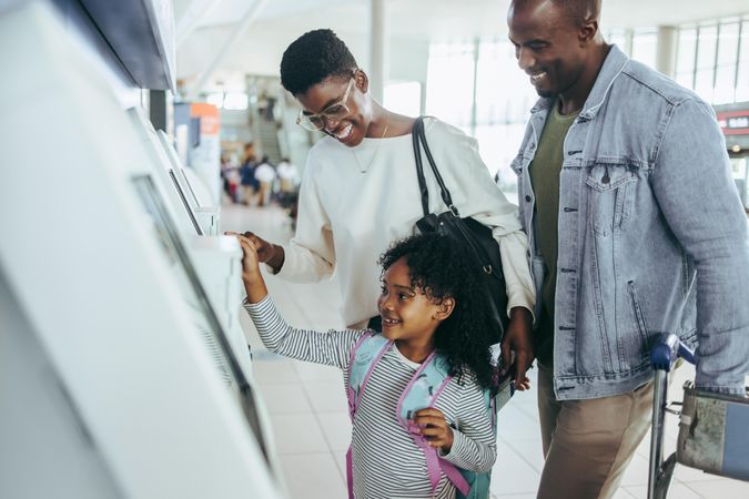 Small girl happy to use self service check in machine at airport with her parents